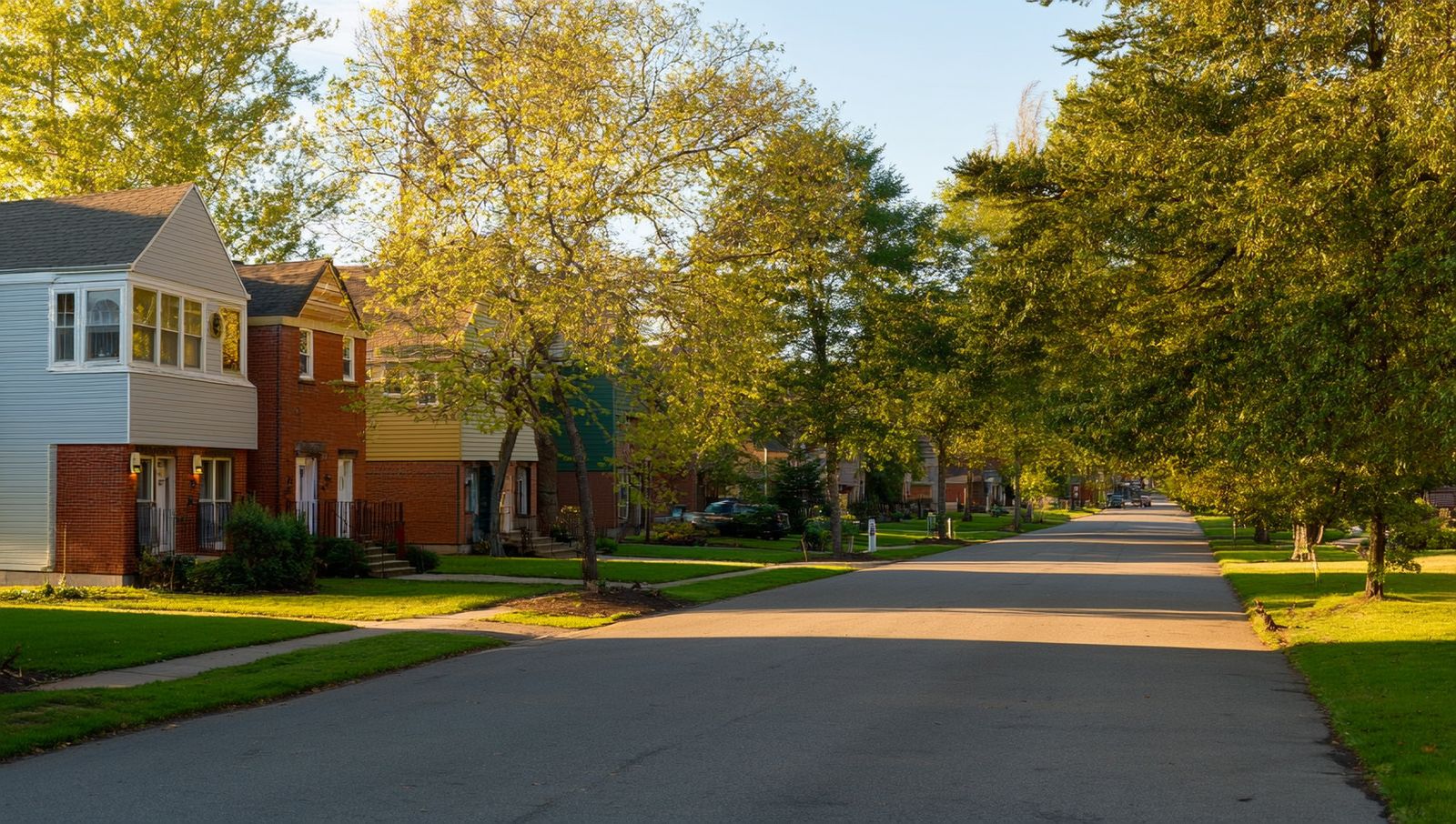A quiet Barrie street with detached brick and siding houses, mature trees and empty driveways bathed in afternoon light